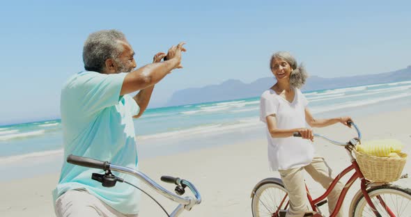 Side view of active senior African American man taking photo of woman with mobile phone at beach 4k