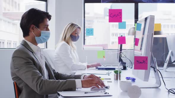 Diverse business colleagues wearing face masks working at desks in office with hygiene screen betwee