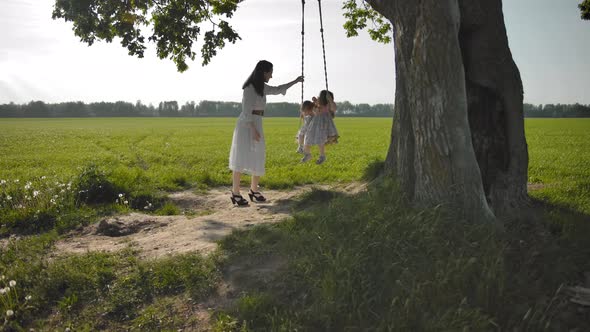 Young Mother Gently Shakes Her Two Daughters 1 and 3 Years Old on a Swing