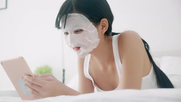Beautiful young asian woman with sheet facial mask lying working on laptop computer on bed.