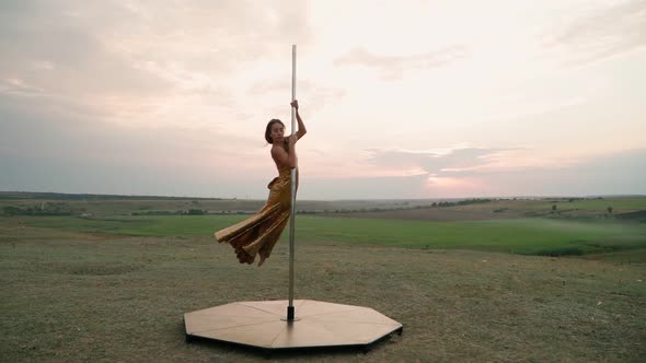 Athletic Woman in a Gold Dress Performs Tricks on the Pole Against the Background of Sunset in Field