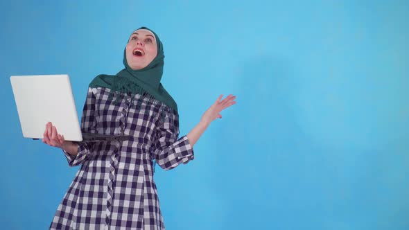 Joyful Young Muslim Woman Uses a Laptop and Learns About the Win on a Blue Background