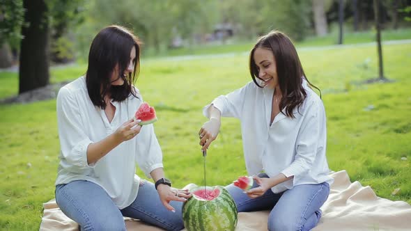 Two Cheerful Brunettes Enjoy Summer Vacation Eating Delicious Watermelon Sitting on Grass in the