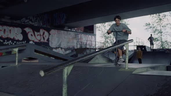 Millennial Man Jumping on Rail with Scooter at Urban City Skate Park