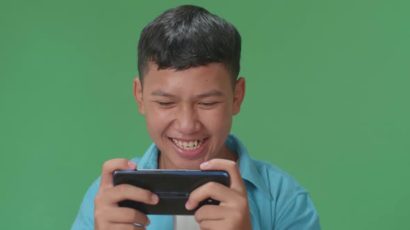 Excited Young Asian Boy Playing Video Game With Mobile Phone In Green Screen Studio alt