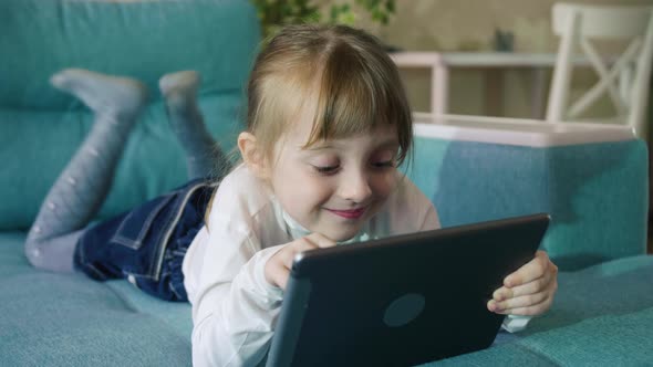 Adorable Kid Girl Using Digital Tablet Watching Cartoons Lying on Bed at Home