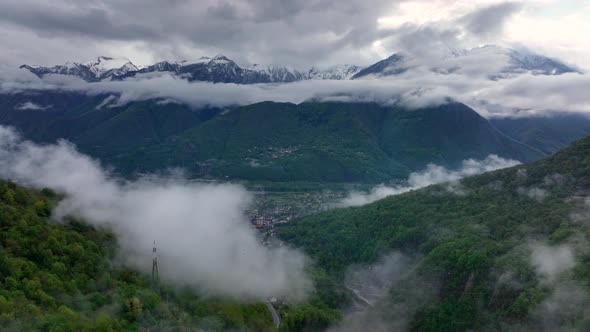 Low clouds over Italian alp village in lush valley by pointed mountains - drone