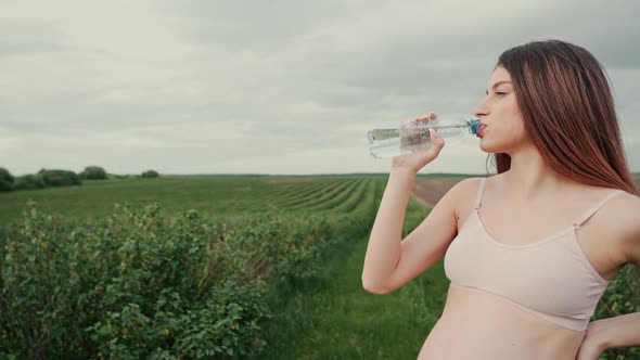 Pregnant young woman with dark hair in a pink top and light pants drinks water outdoors. 