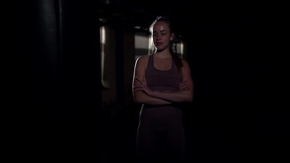 Young Confident Woman with Crossed Hands Standing in Darkness at Punching Bag Smiling Looking Away