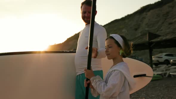 Surfing. Father with sup board and daughter with paddle are walking along the beach