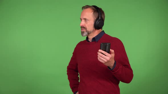 A Middleaged Man Listens to Music with Headphones on and a Smartphone and Smiles  Green Screen