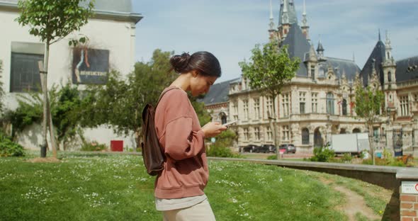 A Beautiful Young Woman Uses a Smartphone While Walking Around a European City