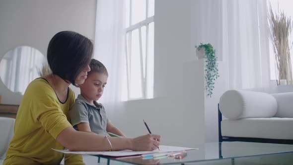 Mother in Yellow Jacket and Son in Tshirt Sit at the Table and Draw Together Color Pencils on Paper