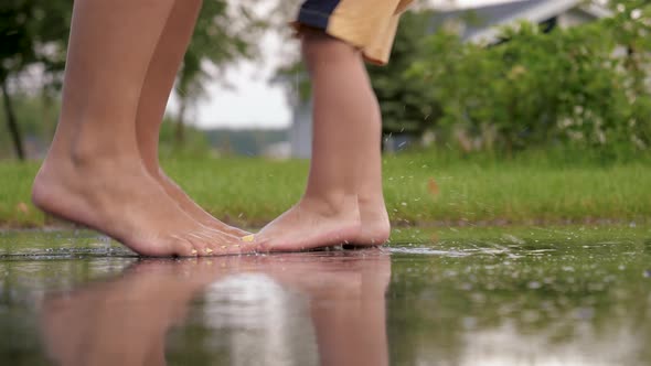 Close Up Legs Of Playful Baby And Mother Barefoot Stomping On Puddle On Asphalt