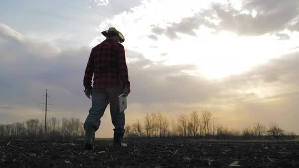 Senior Male Farmer Walks Through a Field with Plowed Land in His Rubber Boots