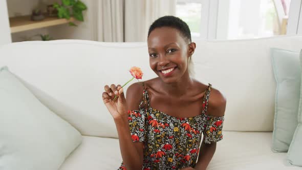 African american woman sitting on sofa smelling flower and smiling to camera