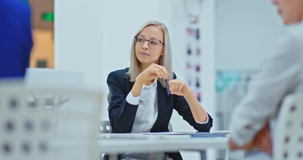 Beautiful Business Woman in Glasses and a Business Suit Sits at the Table and Listens to Her Partner