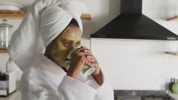 Happy african american woman with beauty mask on face, drinking coffee in kitchen