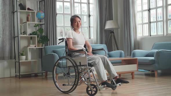 Asian Woman Sitting In A Wheelchair Smiling And Looking At Camera In Living Room