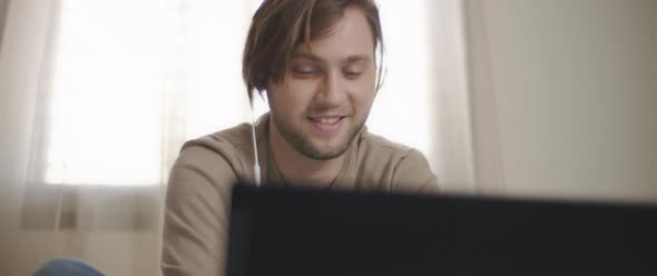 A young man speaking to someone through earphones while working on his laptop. 