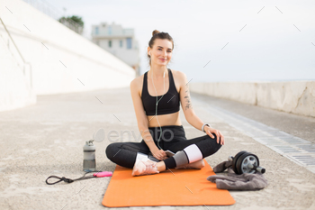 h earphones holding cellphone in hand sitting in lotus pose on orange yoga mat happily looking in camera outdoor