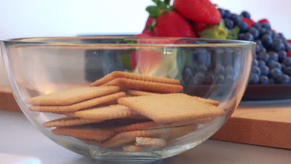 A Woman Puts Cookies Into a Glass Bowl, a Plate with Fruit in the Background - Closeup on the Bowl