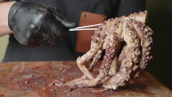 A male chef wearing black gloves, hold up a half cooked octopus with a tong from a chopping board, s