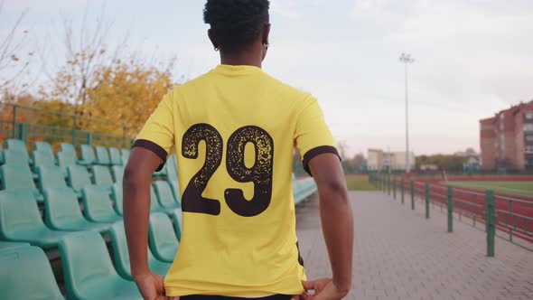 A Young AfricanAmerican Woman Stands in a Soccer Jersey at the Stadium and Points with Two Thumbs to