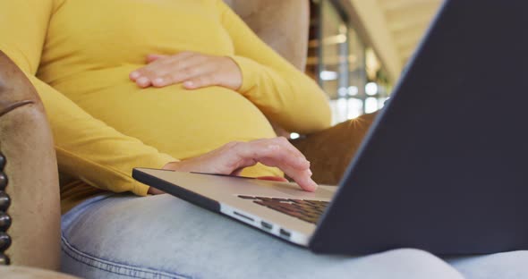 Midsection of caucasian pregnant woman sitting in armchair with laptop