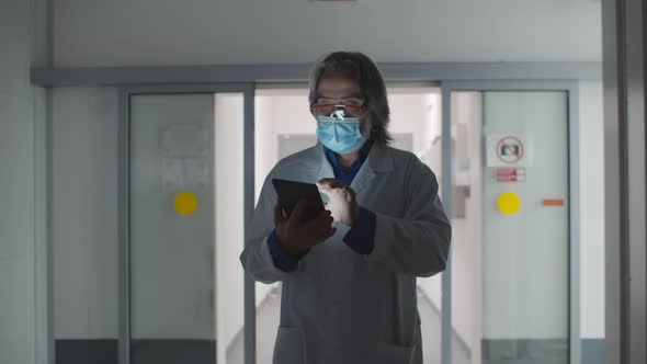 Senior Male Doctor Using Tablet Computer Walking in Hospital Corridor