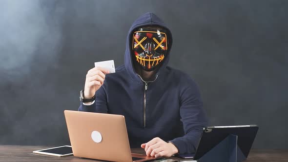 Portrait of a Masked Male Hacker with a Laptop and Credit Card in Hand.