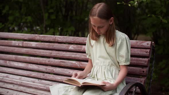 Teenager Girl Leafing Through Book Sitting on Park Bench at Summer Weekend