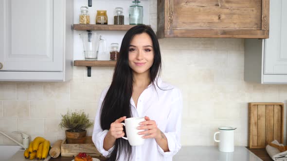 Young Woman Smiling Into Camera With Cup Of Coffee In Her Kitchen