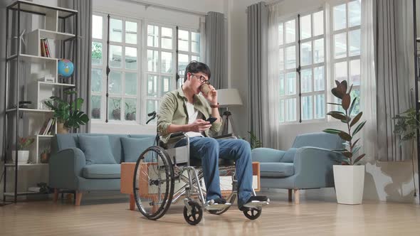 Asian Man Sitting In A Wheelchair While Use Mobile Phone And Drinking Coffee At Home