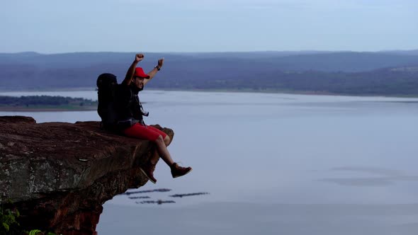 cheerful hiker man sitting and gesture raised arms on the edge of cliff