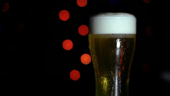 A Glass of Cold Beer on a Black Background with Colored Lights is on the Bar