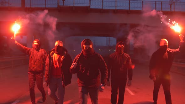 Group of Young Men in Balaclavas with Red Burning Signal Flare Walking on the Road Under the Bridge