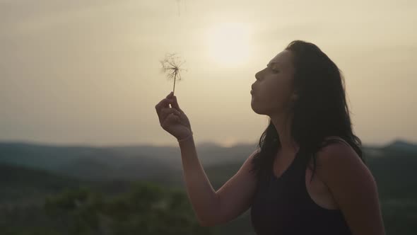 Beautiful Girl Blowing on a Dandelion in the Mountains at Sunset