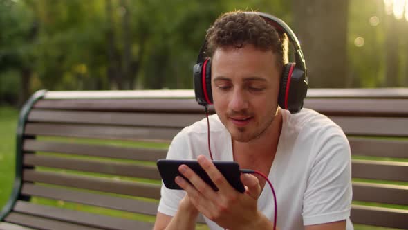 A Young Handsome Guy in a White Tshirt and Headphones Sits on a Bench in the Park at Sunset and