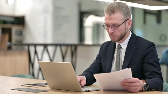 Serious Businessman Working on Laptop with Documents in Office 