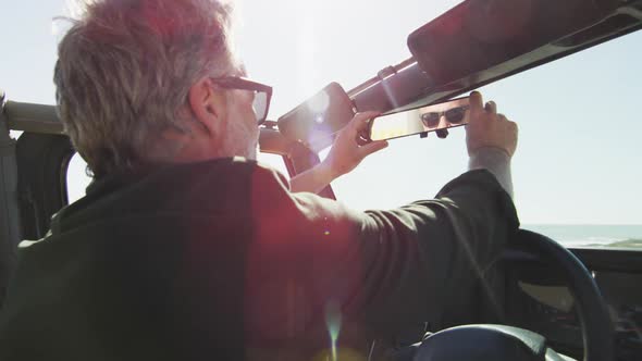 Caucasian man in sunglasses sitting in car adjusting rearview mirror on sunny day at the beach