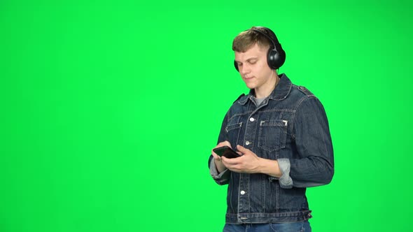 Man in Big Headphones Goes and Texting with Smartphone on Green Screen at Studio.