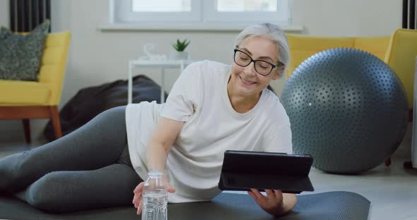 Old Woman in Glasses in Home Clothes Resting on Mat at Home and Watching Interesting videos