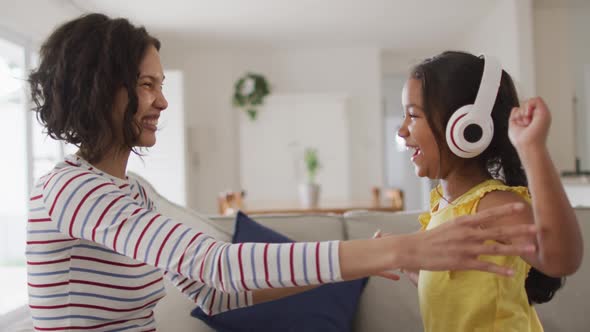 Happy hispanic mother and daughter wearing headphones listening to music sitting on sofa