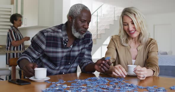 African american senior man and caucasian senior woman sitting by table doing puzzles drinking tea
