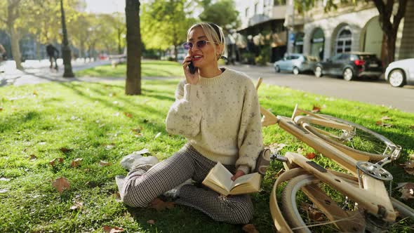 Woman Talking on Smartphone While Reading Book Near Bicycle