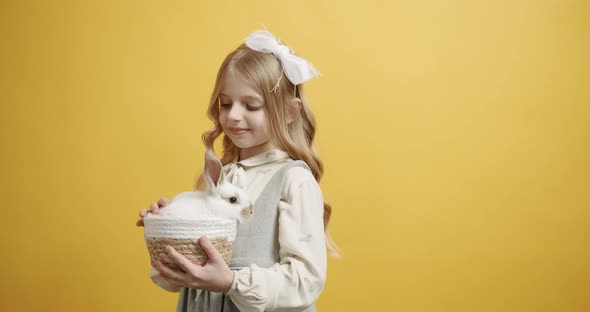 Little Girl Smiles in a White Dress Holds a Basket with a Rabbit in Her Hands