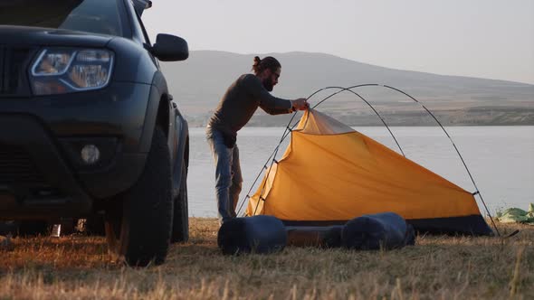 Young Bearded Man Sets Up a Tent at Campsite on the Lake Shore Near His Car. 