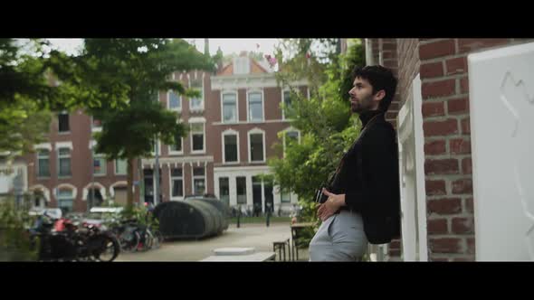 Caucasian man with beard and photocamera leaning up against a building in the city