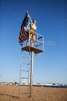 nding on lifeguard place and raising hand with American flag while celebrating USA federal holiday